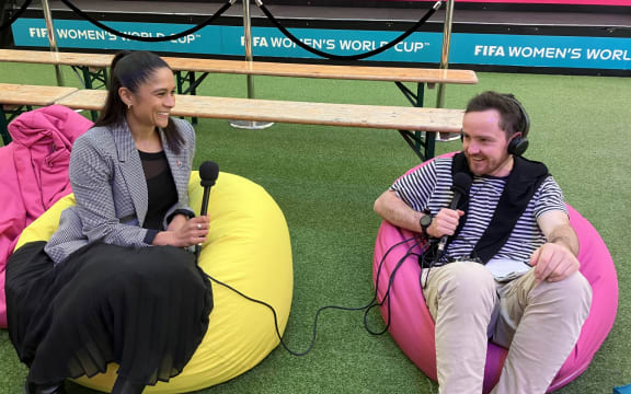 Santha and Tom sit on bean bags holding microphones. They smile as they talk.