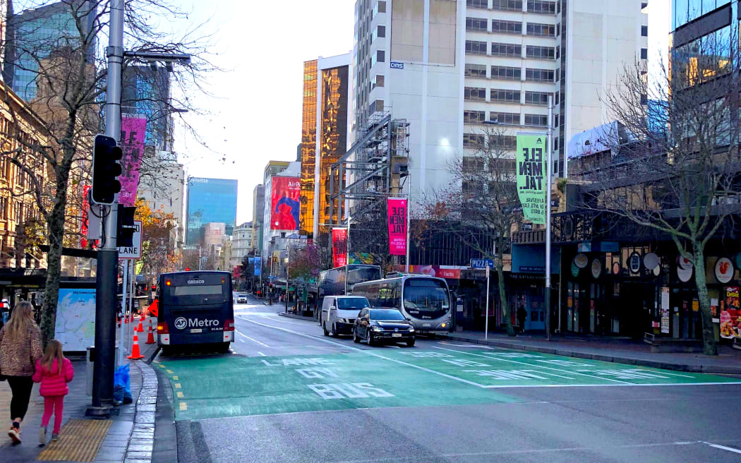 Queen Street on a quiet weekday afternoon. The sky is pale blue, the streets are in shadow. A few buses and cars busy the roads. A woman and her daughter walk along the sidewalk on the left.