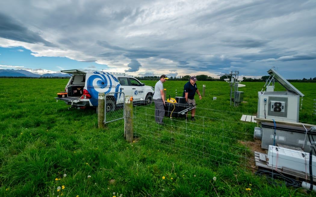 Two individuals carrying scientific equipment in a grassy field. A white vehicle with the "NIWA" logo and a blue swirl is parked nearby, its back door open to reveal more equipment. The individuals are setting up or maintaining instruments within a fenced area containing weather monitoring devices and other technical apparatus. The sky is overcast, and mountains are visible in the distance.