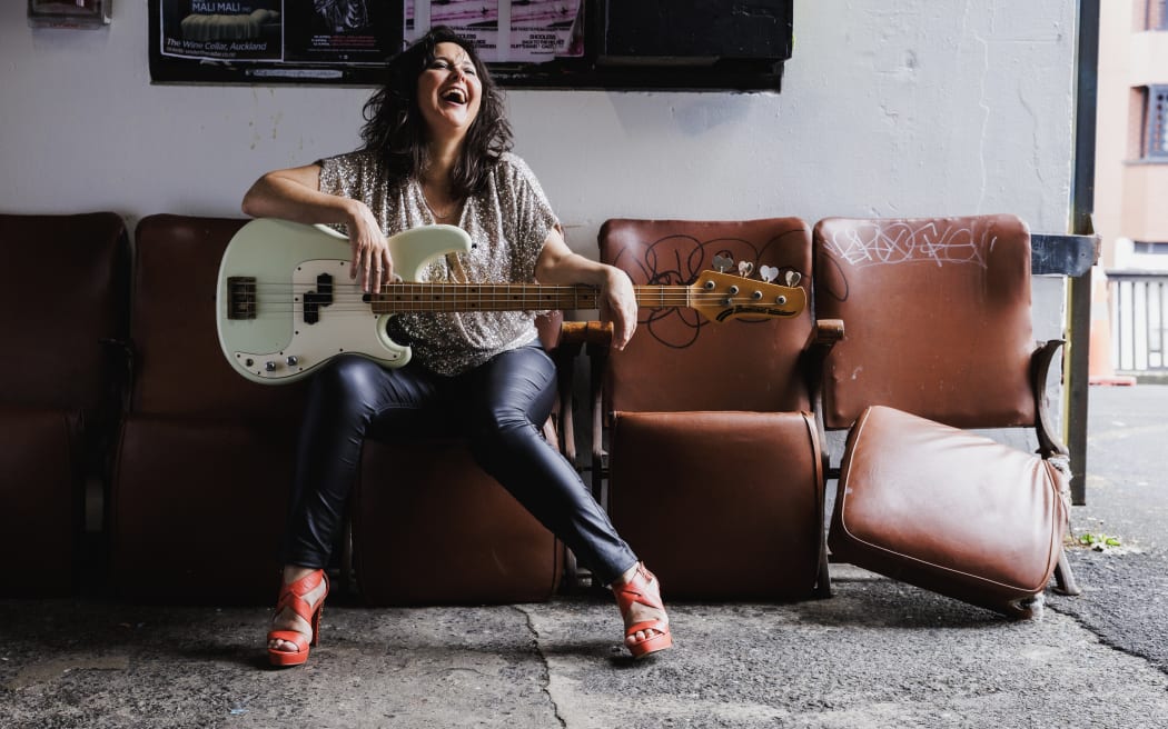 Jodie Rimmer is in character as Nichola Cheesman. She's sitting on a brown lather chair and holdin a bass guitar. She's looking up to the left and laughing.