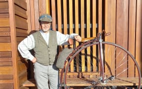A man dressing in Victorian era clothing stands next to his Penny Farthing, with a wooden lined wall in the background.