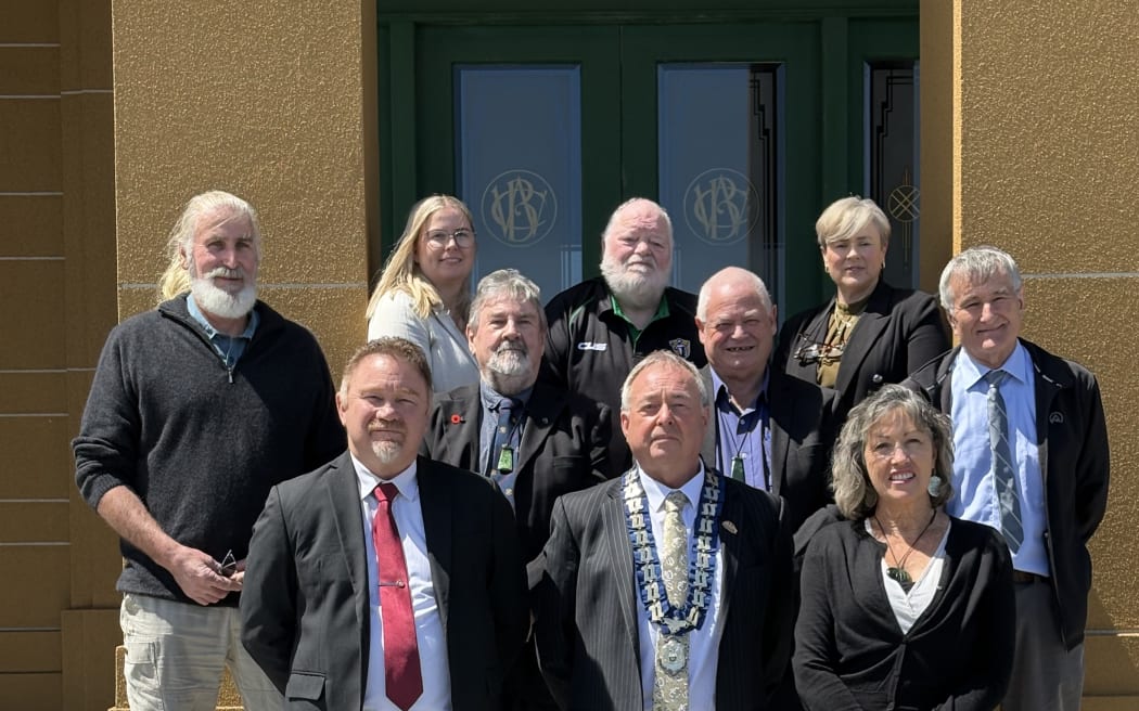 10 councillors, both men and women, stand on steps in the sunshine with a mayor wearing chains of office in centre on bottom step