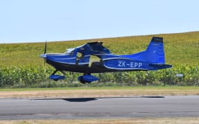 An E-350 plane landing on a runway. It is bright blue, landing in front of a bright green cornfield.