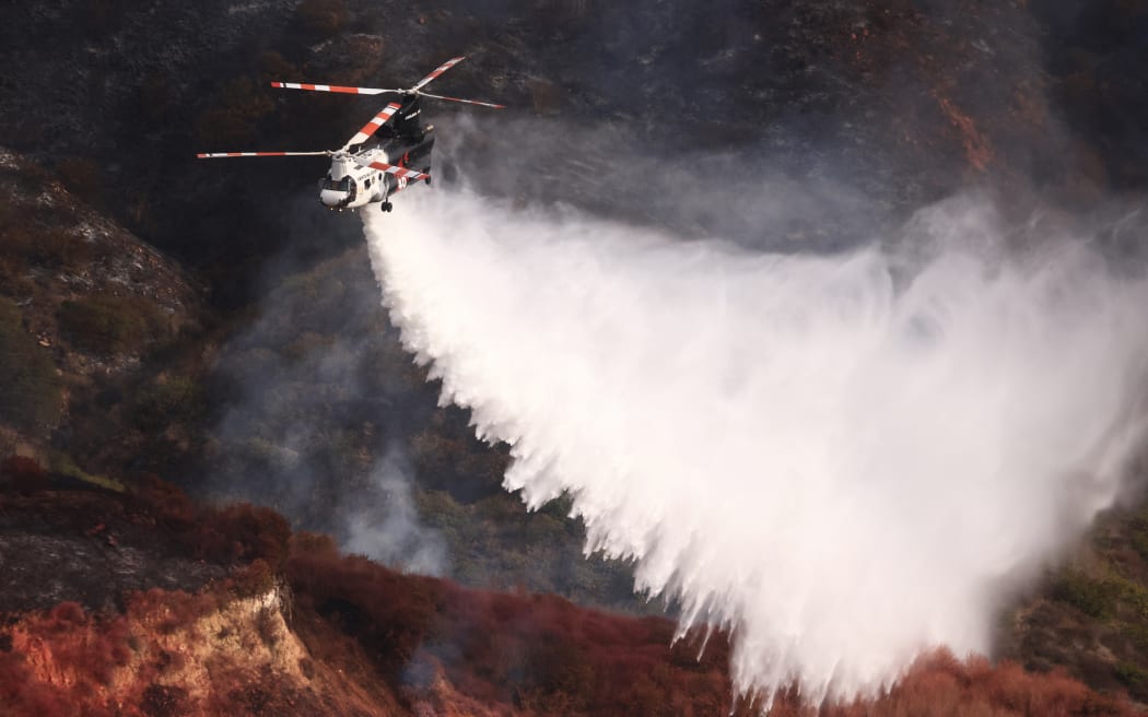 A firefighting helicopter drops water on a mountainside as the Franklin Fire burns in Malibu, California, on December 11, 2024. - The fire which has sent thousands of people fleeing for safety, including Hollywood celebrities, forged west on December 11, as firefighters battled to save homes from the blaze. Around 4,000 acres (1,600 hectares) have been charred by the fire since it erupted late December 9 in the hills above Pepperdine University as the Franklin fire burns around Malibu, California on December 11, 2024. (Photo by DAVID SWANSON / AFP)