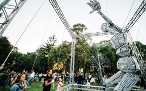 A giant puppet, rigged to a large square scaffold, is puppeted by several people holding long strings.