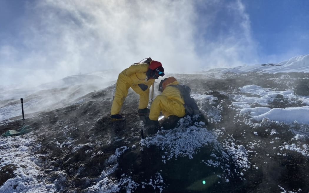 Two people in bright yellow hazmat suits, beanies and eye goggles crouch on the dark volcanic soil strewn with snow on a slope. Volcanic steam rises around them into a bluebird sky.