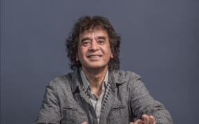 Zakir Hussain sitting cross-legged playing the tabla in grey clothing.
