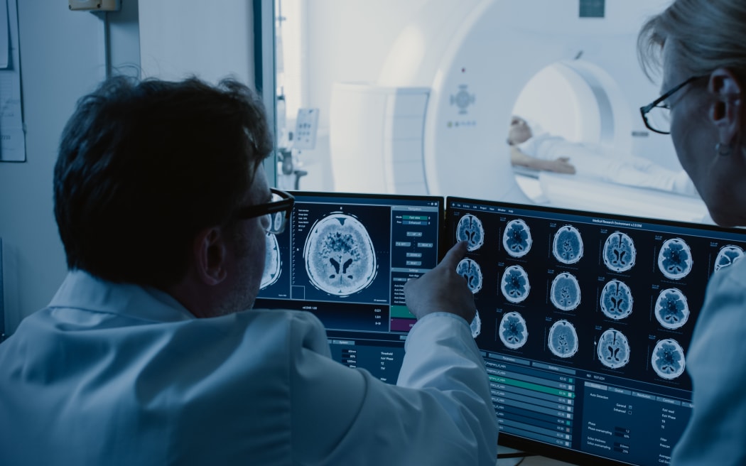 In Control Room Doctor and Radiologist Discuss Diagnosis while Watching Procedure and Monitors Showing Brain Scans Results, In the Background Patient Undergoes MRI or CT Scan Procedure.