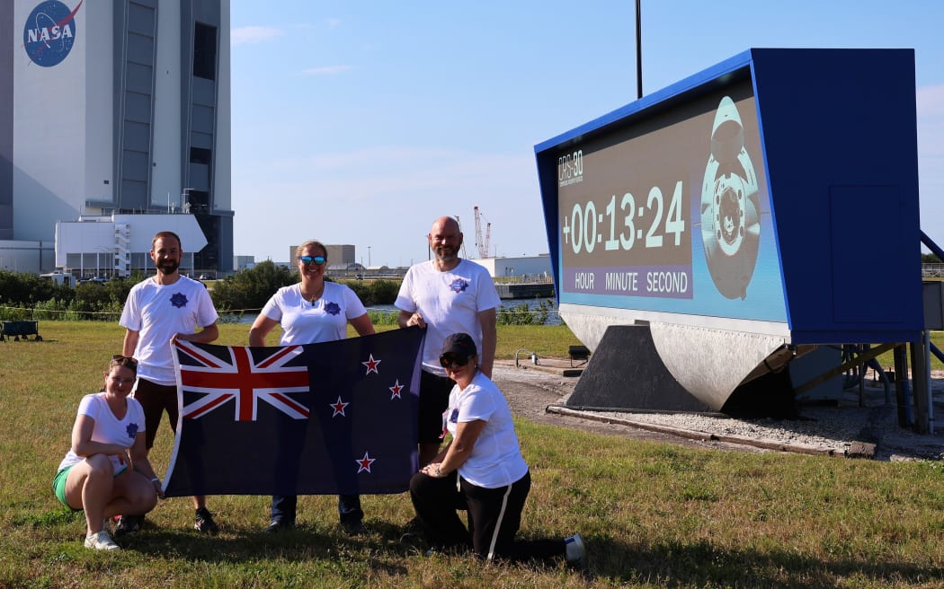 A group of five people wearing white shirts with a logo are posing outdoors in front of a large countdown timer display and holding a New Zealand flag. In the background, there is a tall building with the NASA logo on it.