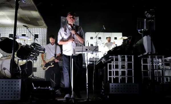 Three men in white shirts and grey pants play instruments on a darkened stage.