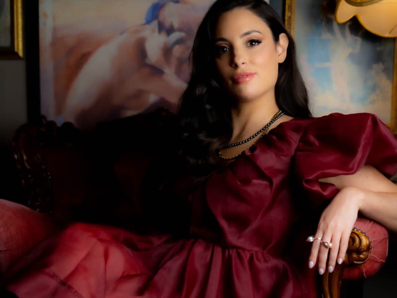 Jade May - a woman with long dark hair and a glamourous deep red gown - lounges in a chair.