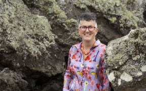 Dr Lucy O'Hagan - a woman with short grey hair and red glasses - stands smiling between some large rocks.