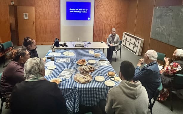 A group of people sit on chairs around a table of food, facing the front of the room. Yvonne Glassborow sits in front of the projector sharing a presentation with the group.