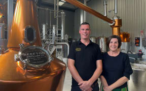 Gareth and Patsy stand smiling in front of brass distilling equipment in a warehouse.