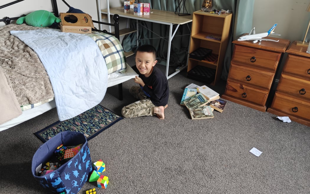 Raymond playing around at home in a room with a bag full of Rubik's cubes, books on the floor and board games in a shelf.