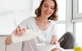 Portrait of a smiling young woman pouring milk into the bowl while having breakfast at the kitchen