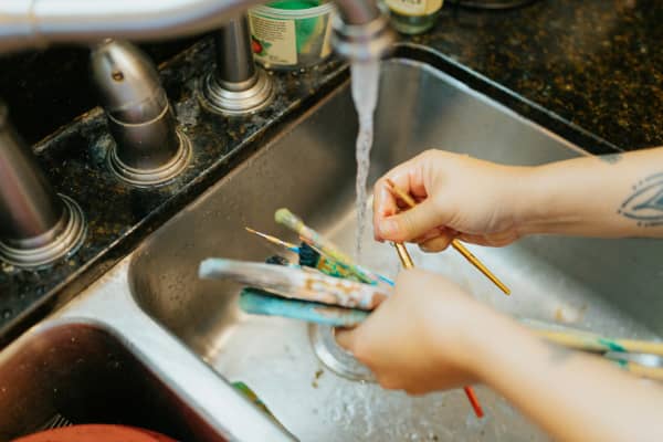 Paintbrushes being cleaned in a sink under running water of tap.