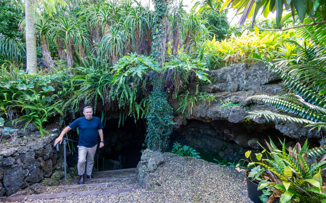 A wide shot of a man in a navy tshirt standing on steps descending into a dark cave. The cave entrance is surrounded with lush ferns and other foliage.