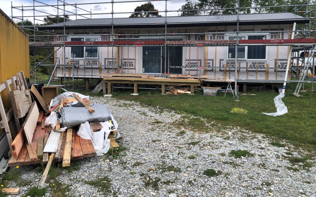 A small house is under construction on a plot of grassy land. There is scaffolding around the structure and the exterior walls are not yet finished. There is a shipping container to the right with building materials strewn around it. It is a cloudy day.