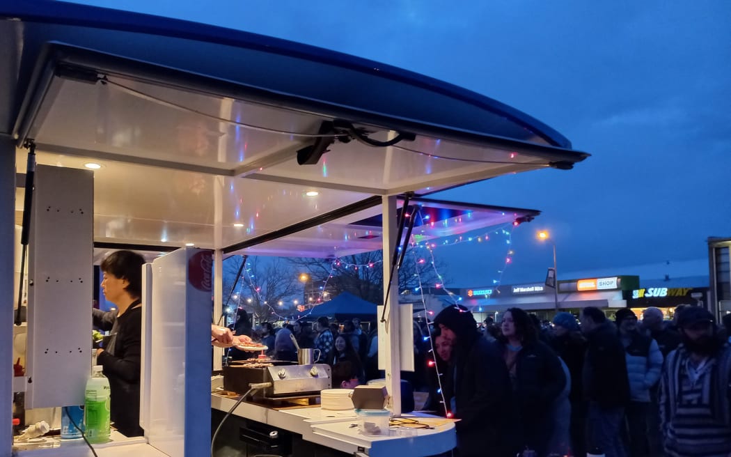 A bustling market at twilight. A food truck strung with fairy lights is busy making orders.