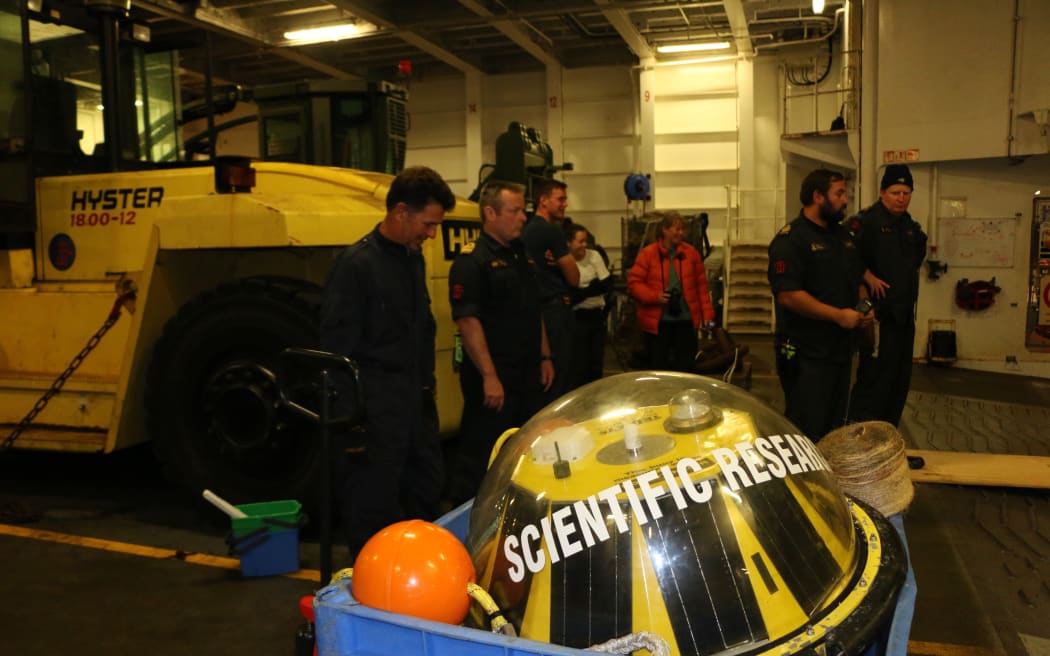 People stand below deck on a ship near a large plastic buoy with the words 'SCIENTIFIC RESEARCH' written on it.