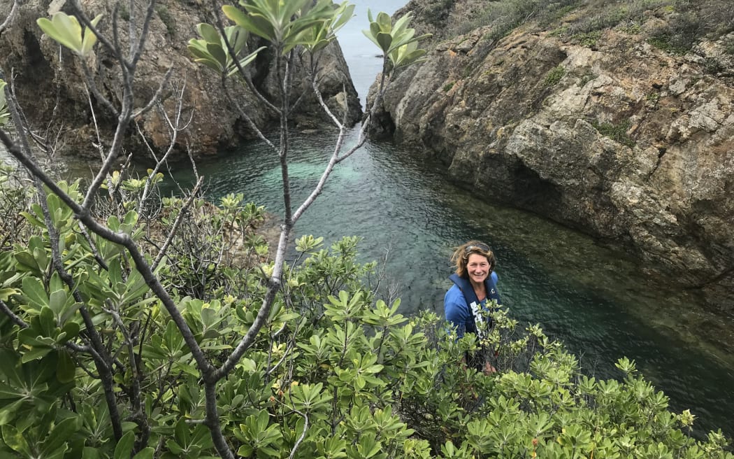 A woman is smiling at the camera from behind a foreground of pōhutukawa with a clear ocean pool between two large rocks behind her.