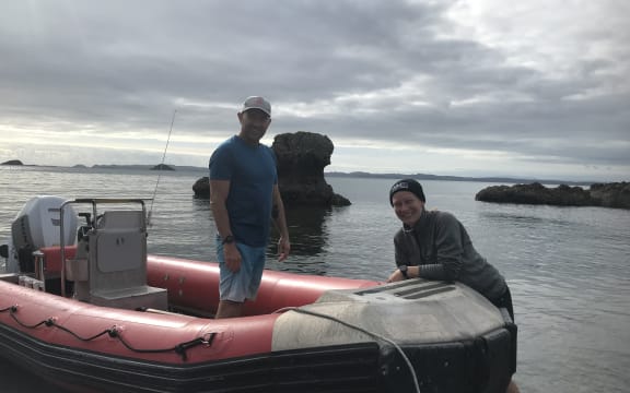 A man stands in an inflatable zodiac boat pulled ashore in the shallows while a woman stands next to the boat. The pair are looking at the camera and smiling.