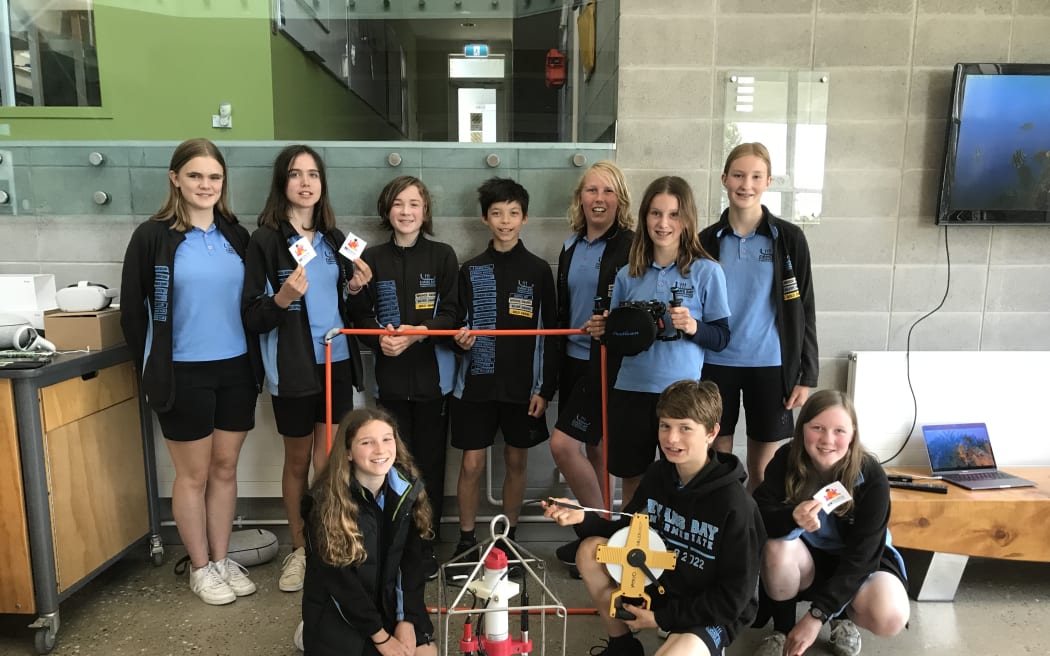 A group picture of the Evans Bay Intermediate students group. They are holding marine biology equipment - a quadrat, a piece of CTD equipment, a transect tape and a small orange ROV.