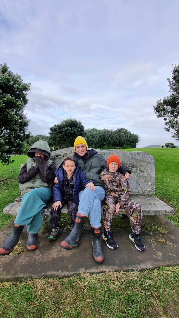 Kara Rickard, wearing gumboots and a yellow beanie, sits on a park bench with three kids wearing warm clothes.