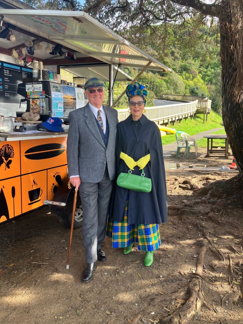 Jacqui and her husband stand outside a food truck in their vintage clothes. Jacqui wears a navy cape, blue and yellow tartan skirt, yellow gloves, a green bag, and a matching tartan bonnet.