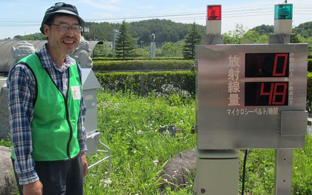 Japanese man in fluoro vest stands beside stainless steel box with digital display