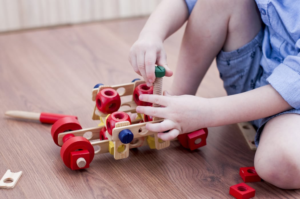 the little boy twists the bolt into a wooden machine  on floor in room.