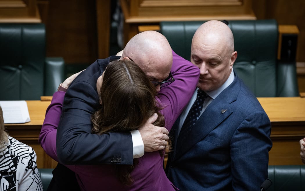 Nicola Willis elbows Christopher Luxon in the face while embracing Todd Muller after his valedictory address.