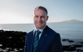 Climate Change minister Simon Watts, wearing a blue suit, stands in bright sunlight near a rocky shore, with Rangitoto Island in the background.