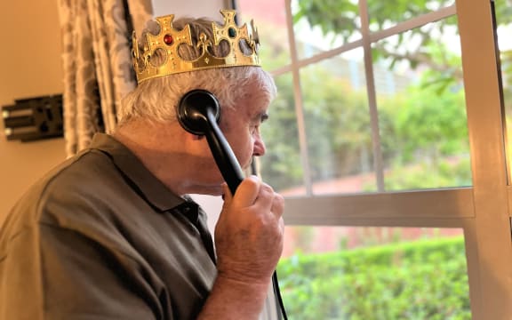 Tom, an elderly man in a brown t-shirt, looks out a window wearing a golden crown and holding an old-fashioned black rotary phone.