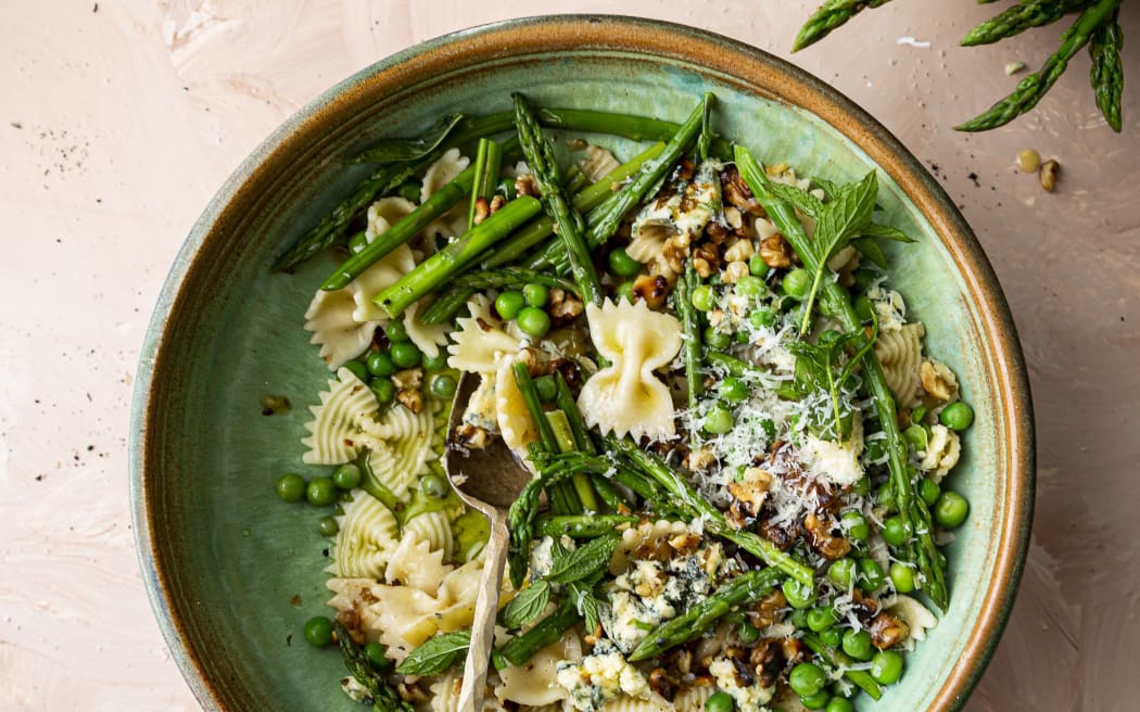 A bowl of farfalle (bow-shaped) pasta with asparagus, peas, blue cheese and walnuts on a pale pink background.