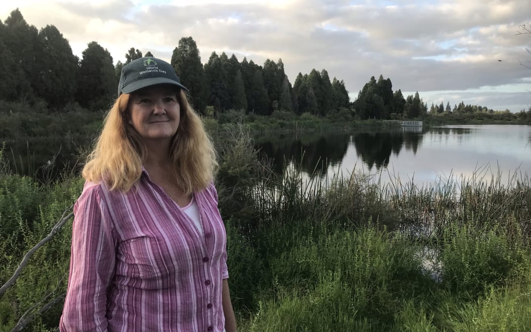 A woman wearing a cap stands in front of a lake and wetland vegetation on a cloudy day.