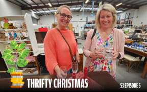 Two smiling women stand inside a thrift store, holding various glassware items. The woman on the left wears a bright orange sweater, and the woman on the right wears a floral dress with a light pink cardigan. The background features shelves filled with second-hand items, including stuffed toys and clothing. A money plant graphic sits in the lower-left corner, and the text reads 'Thrifty Christmas S3 Episode 5.