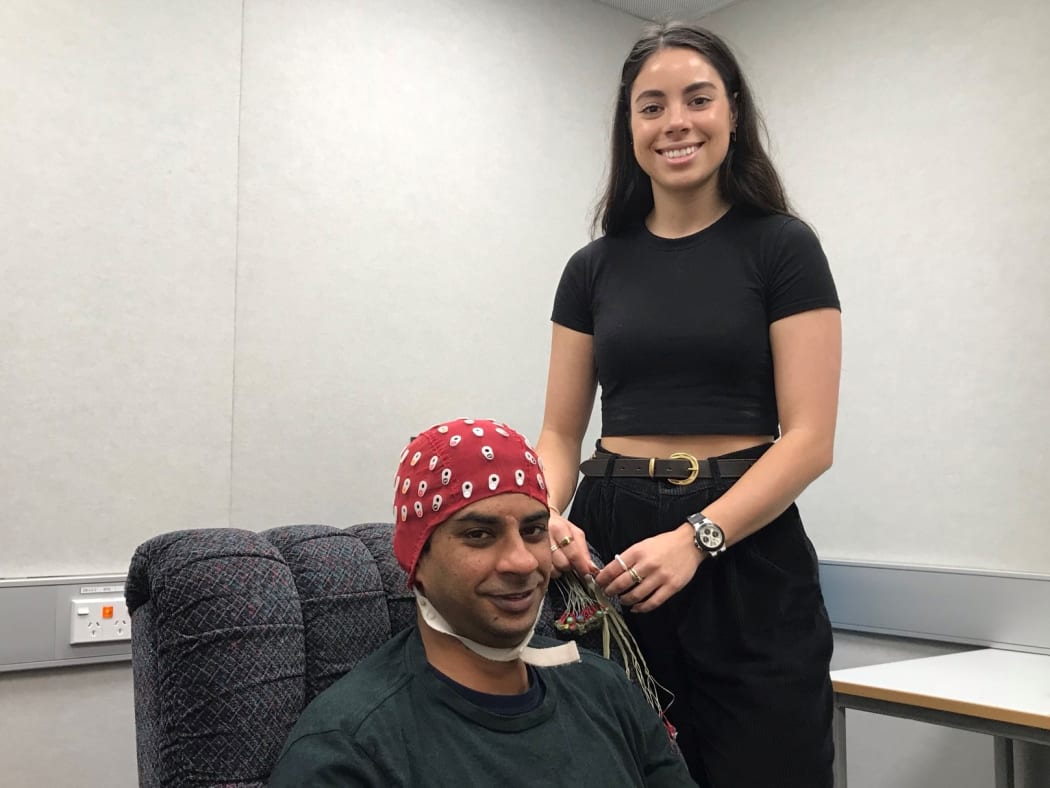 Dunja is smiling, wearing a black crop top and jeans, holding a bundle of wires. Beside her Amit sits in a comfy chair, with the red EEG cap on his head, you can see the labelled holes where the wires will be attached.