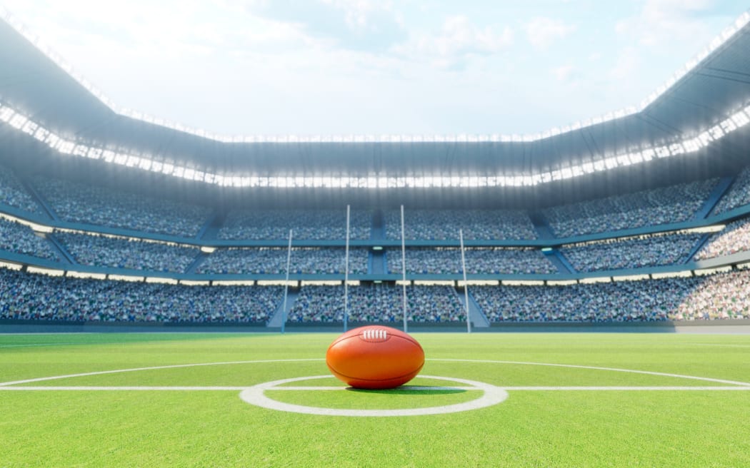A red Australian Rules ball on the centre line of a stadium with posts on a marked green grass pitch in the day time.