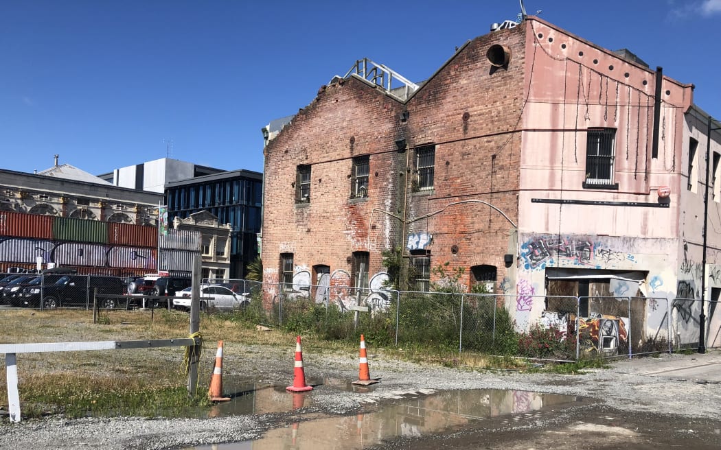 A damaged brick building in central Christchurch, next to an empty site with a pool of water and orange cones.