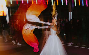 A person in a white dress is dancing in the street waving some orange scarves. It is early evening. There is someone roller skating in the background, and a crowd of people watching.