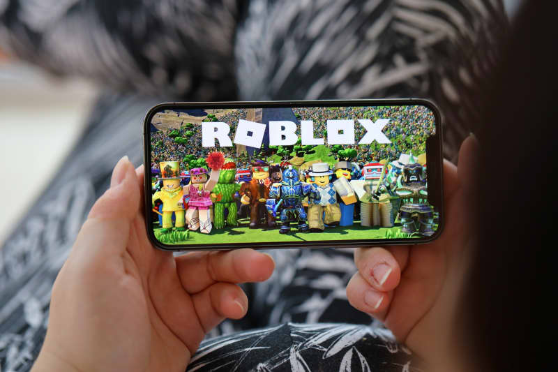 A pair of hands hold an iPhone 15 showing the Roblox mobile iOS game.