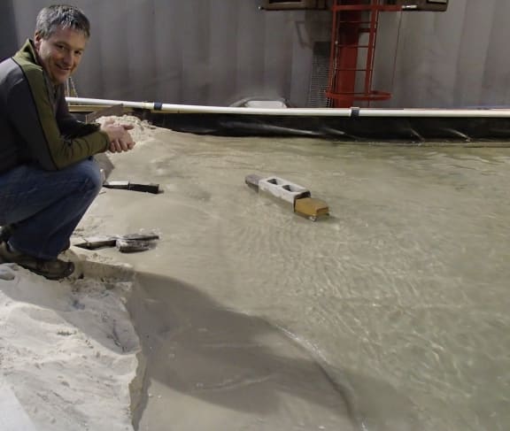 Crile Doscher with the 1:50 scale model of Wairewa/Lake Forsyth. He is crouching on Kaitorete Spit, the outlook to the lake is to his left, and the bricks are simulating breakwaters.