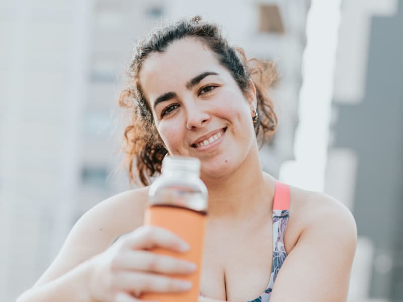 A smiling woman with brown hair pulled into a pony tail wears workout gear and holds a water bottle.