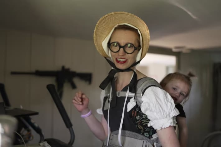 Simone Collins, wearing a pilgrim-style dress topped off with a bonnet, carrying one of her children on her back