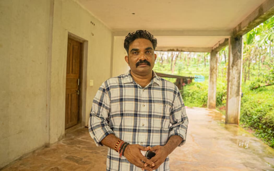 An Indian man with a black moustache wearing a black and white plaid shirt standing under a concrete balcony.