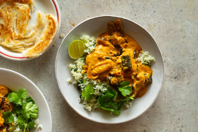 A bowl of yellow-orange toned eggplant curry on rice, with coriander leaves and lime cheeks, with a bowl of roti in shot.
