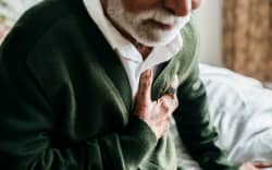 An elderly man sitting down leans forwards as he holds his chest.