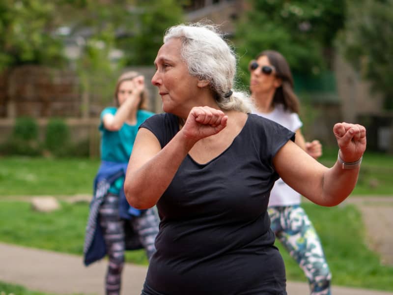 A white-haired woman in a black t-shirt raises her arms in an outdoor exercise class.